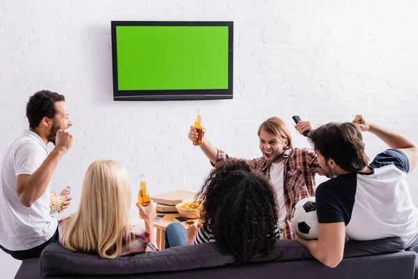 Excited multicultural football fans showing success gesture and holding beer near led tv on wall — Stock Photo