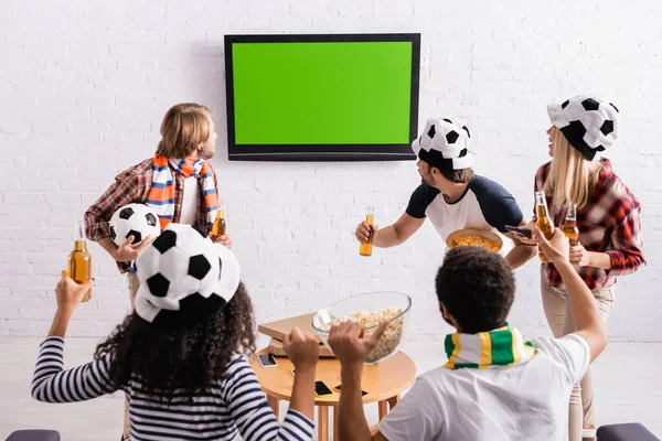 Multicultural friends in football fans hats watching championship on tv — Stock Photo