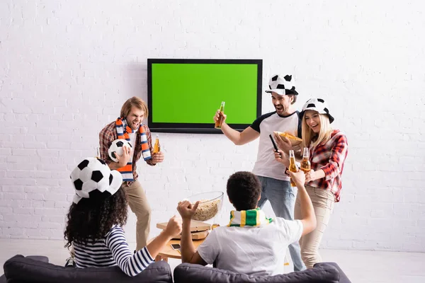 Весёлые мультикультурные друзья в шляпах футбольных болельщиков, держащие пиво возле ЖК-телевизора на стене — стоковое фото
