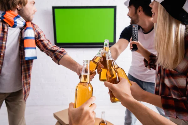 Multiétnicos fãs de esportes clinking garrafas de cerveja enquanto assiste competição desportiva na tv no fundo turvo — Fotografia de Stock