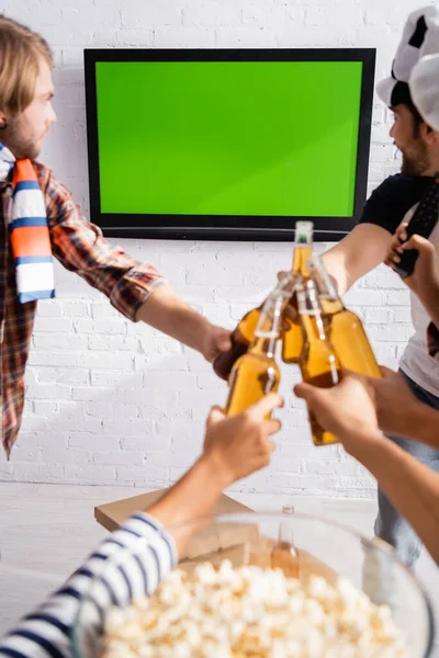 Multiethnische Fußballfans klirren mit Bierflaschen, während sie die Meisterschaft im Fernsehen in der Nähe von Popcorn auf verschwommenem Hintergrund verfolgen — Stockfoto