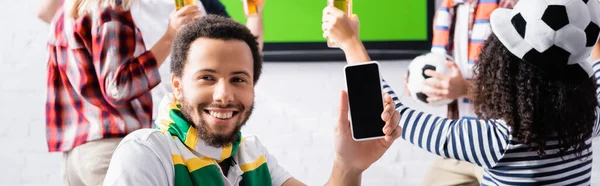 Homme afro-américain joyeux tenant smartphone avec écran blanc près des fans de football multiethnique sur fond flou, bannière — Photo de stock