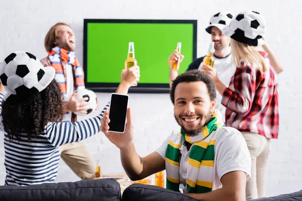 Fröhlicher afrikanisch-amerikanischer Mann zeigt Smartphone mit leerem Bildschirm neben multikulturellen Freunden in Fußballfans-Hüten auf verschwommenem Hintergrund — Stockfoto