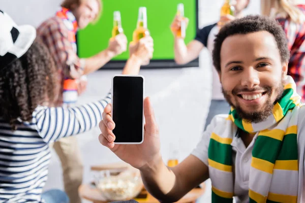 Alegre afroamericano hombre sosteniendo teléfono inteligente con pantalla en blanco cerca de amigos multiculturales en fondo borroso - foto de stock