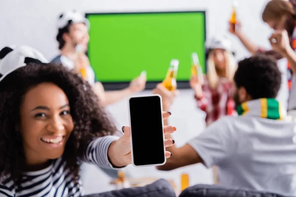 Щаслива афроамериканка, що показує смартфон з порожнім екраном біля мультикультурних футбольних фанатів на розмитому фоні — стокове фото