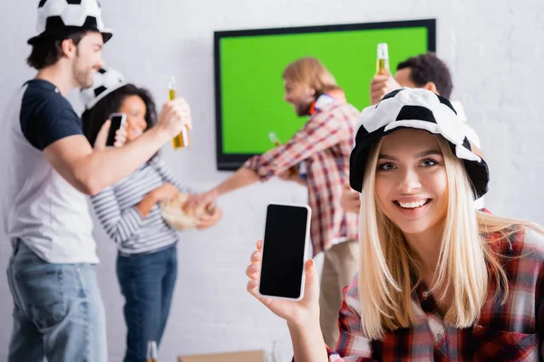 Smiling woman holding smartphone with blank screen near excited multicultural football fans on blurred background — Stock Photo