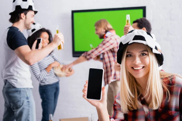 Mujer sonriente sosteniendo teléfono inteligente con pantalla en blanco cerca de aficionados al fútbol multicultural emocionados en un fondo borroso - foto de stock