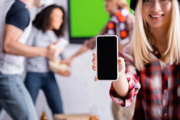 Mulher sorrindo mostrando smartphone com tela em branco perto de fãs de esportes multiétnicos em fundo borrado — Fotografia de Stock