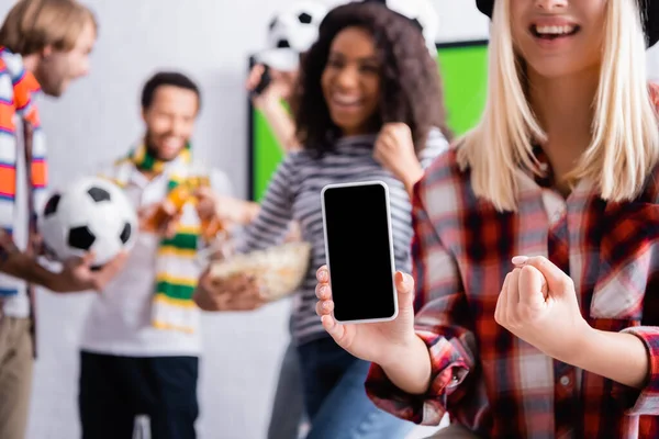 Улыбающаяся женщина показывает победный жест, держа смартфон с пустым экраном рядом с мультикультурными друзьями на размытом фоне — стоковое фото