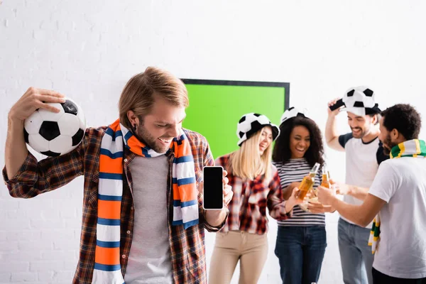 Fröhlicher Mann, der Fußballball und Smartphone mit leerem Bildschirm in der Nähe multiethnischer Freunde hält und auf verschwommenem Hintergrund mit Bierflaschen klingelt — Stockfoto