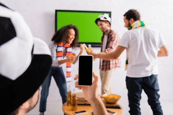 Hombre en fútbol abanico sombrero tomando foto de amigos multiculturales tintineo botellas de cerveza sobre fondo borroso - foto de stock