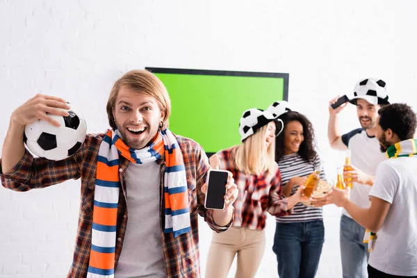 Homme excité tenant ballon de football et smartphone avec écran blanc près d'amis multiethniques sur fond flou — Photo de stock