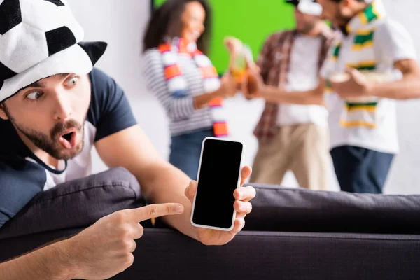 Schockierter Mann zeigt mit Finger auf Smartphone mit leerem Bildschirm in der Nähe multiethnischer Freunde auf verschwommenem Hintergrund — Stockfoto