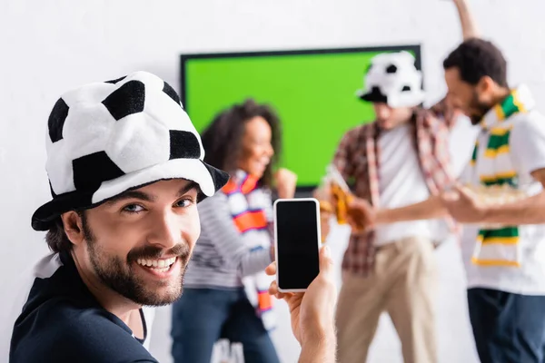 Fröhlicher Mann mit Fußballfanhut zeigt Smartphone mit leerem Bildschirm in der Nähe multiethnischer Freunde auf verschwommenem Hintergrund — Stockfoto