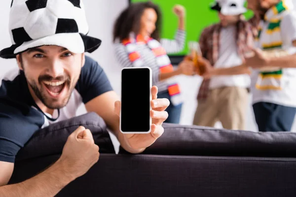 Excited football fan showing win gesture while holding smartphone with blank screen near multiethnic friends on blurred background — Stock Photo