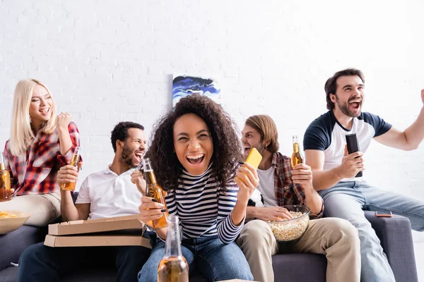 Fröhliche afrikanisch-amerikanische Frau hält Smartphone und Bier in der Hand, während sie mit aufgeregten multikulturellen Freunden Fußballweltmeisterschaft guckt — Stockfoto