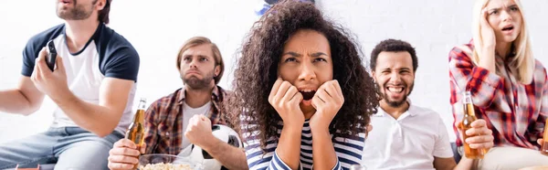 Tendue femme afro-américaine regarder le championnat de football avec des amis multiculturels inquiets sur fond flou, bannière — Photo de stock