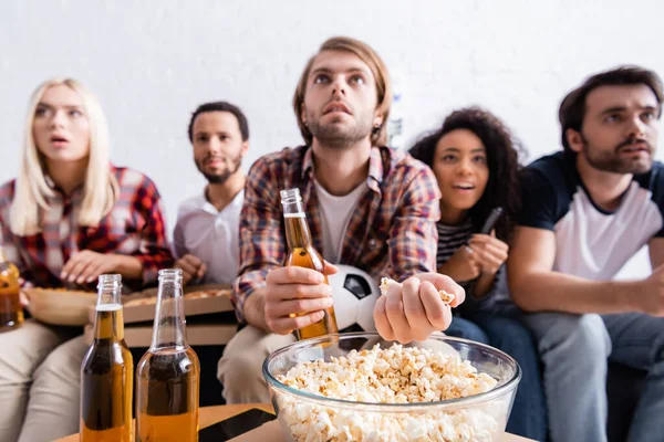 Konzentrierter Mann, der Popcorn trinkt, während er Fußballwettkämpfe in der Nähe multiethnischer Freunde auf verschwommenem Hintergrund verfolgt — Stockfoto