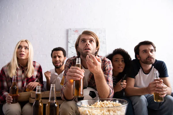 Тривожний чоловік жестикулює, тримаючи пиво і спостерігаючи за футбольним чемпіонатом з багатоетнічними друзями — стокове фото