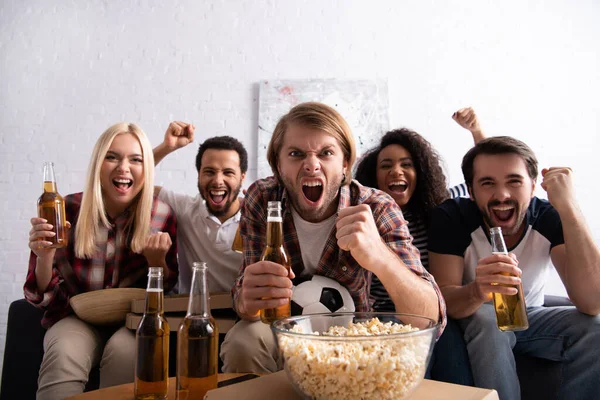 Homme excité crier et montrer geste de victoire tout en regardant le match de football avec des amis multiethniques — Photo de stock