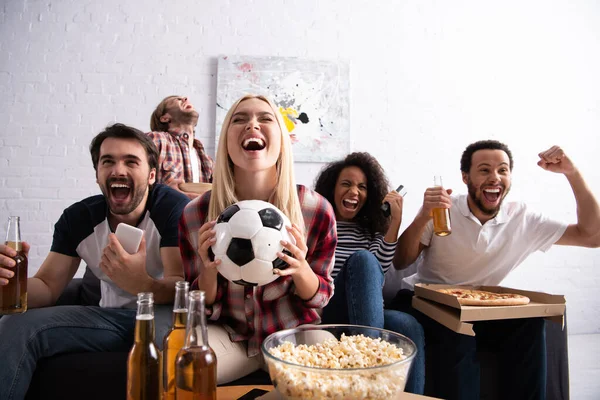 Laughing woman holding soccer ball while watching championship with excited multiethnic friends — Stock Photo