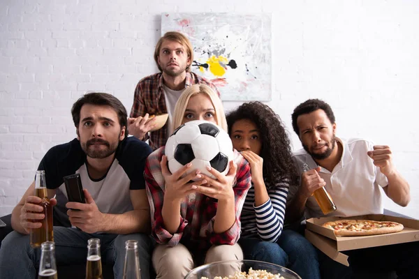 Tense woman obscuring face with soccer ball while watching football championship with worried multicultural friends — Stock Photo