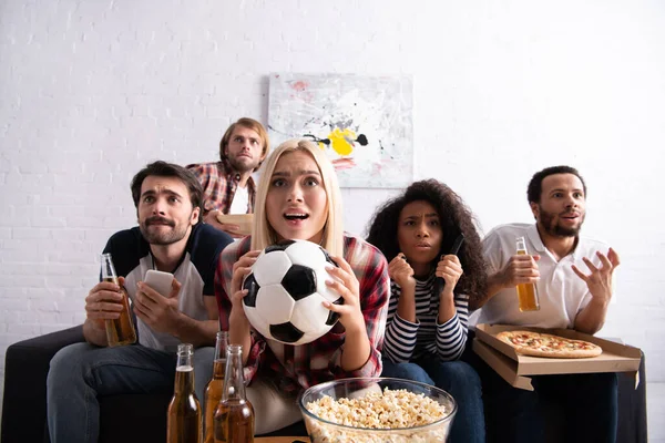 Worried woman holding soccer ball while watching football championship with tense multiethnic friends — Stock Photo