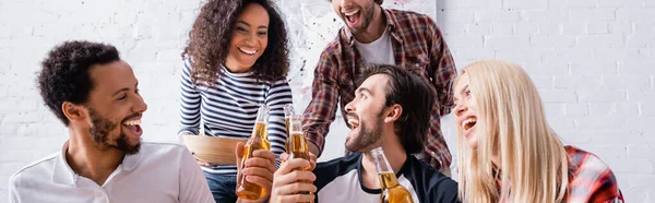 Amigos multiculturales alegres sosteniendo botellas de cerveza durante la fiesta, bandera - foto de stock