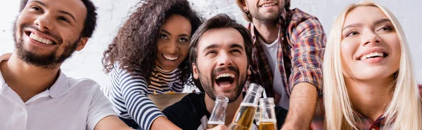 Amici multiculturali eccitati e ridenti che festeggiano a casa e chiudono bottiglie di birra, striscione — Foto stock