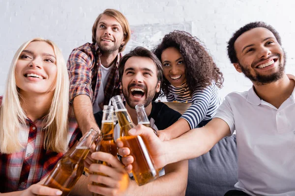 Aufgeregte multiethnische Freunde, die während einer Party mit Bierflaschen im verschwommenen Vordergrund klirren, verschwommener Vordergrund — Stockfoto