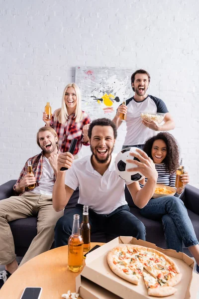 Alegre africano americano hombre celebración fútbol pelota mientras viendo fútbol competencia con emocionado multicultural amigos - foto de stock