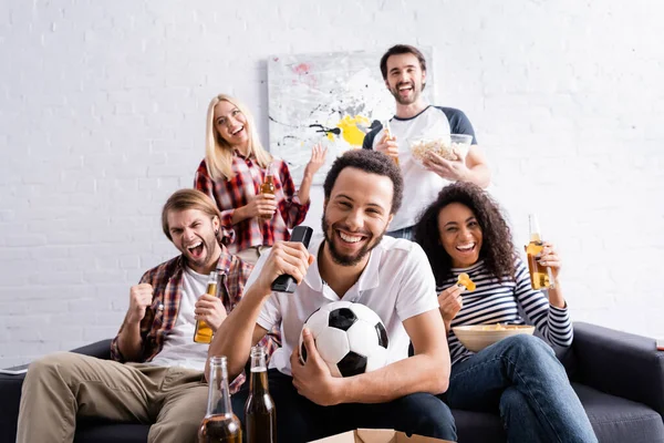 Emocionado hombre afroamericano con pelota de fútbol viendo campeonato de fútbol con amigos multiculturales mostrando gesto de victoria - foto de stock