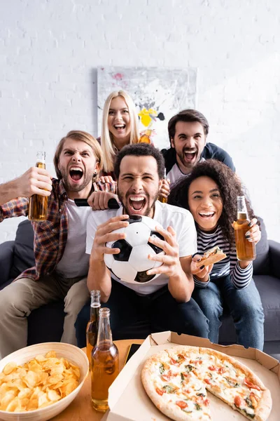 Animado multiétnico fãs de futebol gritando enquanto assiste campeonato perto de cerveja, pizza e batatas fritas — Fotografia de Stock