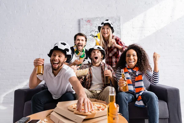 Взволнованный человек, принимающий пиццу во время просмотра чемпионата по футболу с мультикультурными друзьями на размытом переднем плане — стоковое фото
