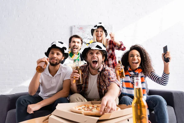 Excited man shouting, holding beer and taking pizza while watching championship with multicultural friends on blurred foreground — Stock Photo