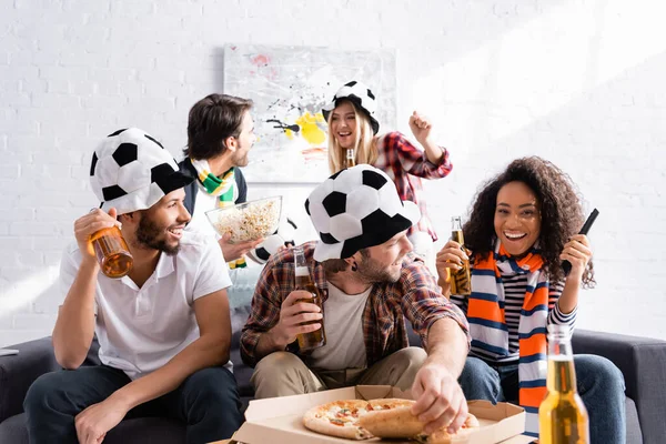 Hombre sosteniendo cerveza y tomando pizza cerca de amigos multiétnicos en los aficionados al fútbol sombreros, borrosa primer plano - foto de stock