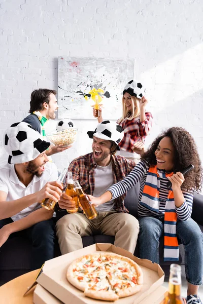 Amigos multiculturais alegres batendo garrafas de cerveja perto de pizza em primeiro plano borrado — Fotografia de Stock