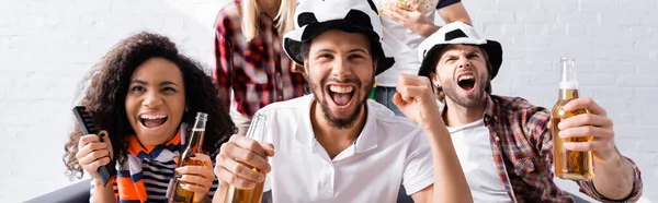 Hombre emocionado gritando y mostrando gesto de victoria mientras ve la competencia de fútbol con amigos multiétnicos en un fondo borroso, pancarta - foto de stock