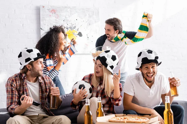 Animado mulher segurando bola e apontando com o dedo perto de amigos multiculturais em fãs de futebol chapéus e scarfs — Fotografia de Stock