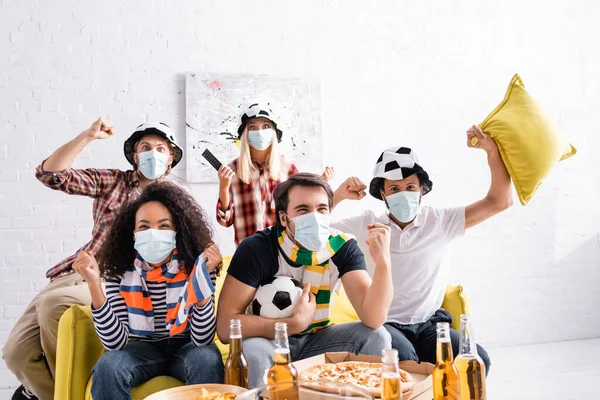 Amigos multiétnicos animados em máscaras médicas e chapéus de fãs de futebol assistindo campeonato em casa — Fotografia de Stock