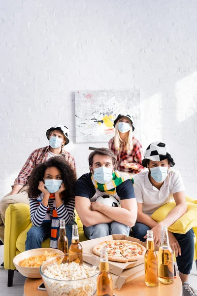 Upset multiethnic friends in medical masks and football fans hats watching championship on blurred foreground — Stock Photo