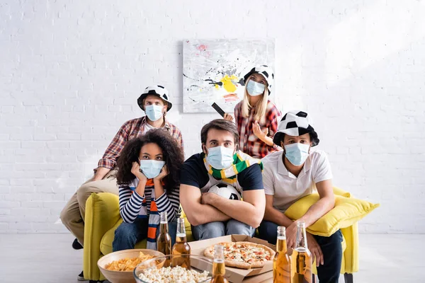 Chateado fãs de futebol multiétnico em máscaras médicas assistindo campeonato em casa — Fotografia de Stock