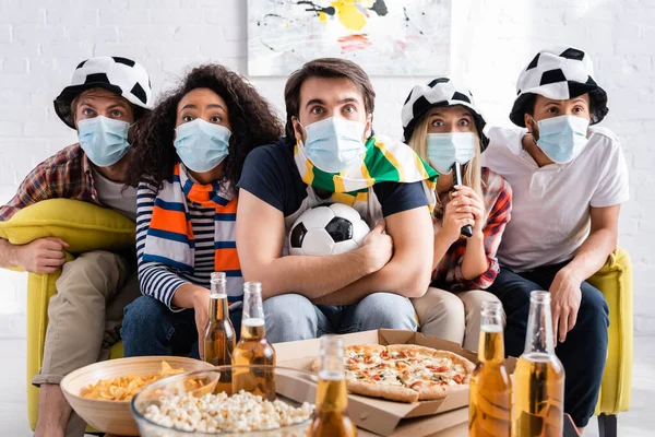 Fanáticos del fútbol multiétnico enfocados en máscaras médicas viendo el partido de fútbol cerca de la cerveza y aperitivos en primer plano borroso - foto de stock