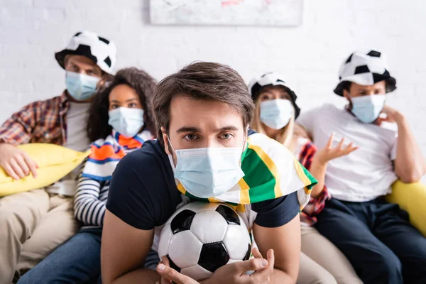 Focado homem em máscara médica segurando bola de futebol enquanto assiste competição de futebol com amigos multiétnicos em fundo turvo — Fotografia de Stock