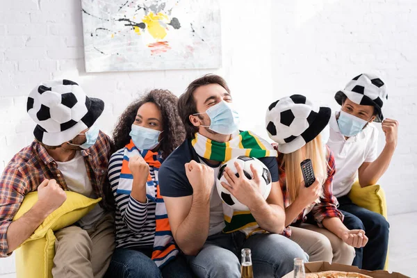 Hombre feliz con los ojos cerrados sosteniendo pelota de fútbol cerca de amigos multiculturales en los aficionados al fútbol sombreros y máscaras médicas - foto de stock