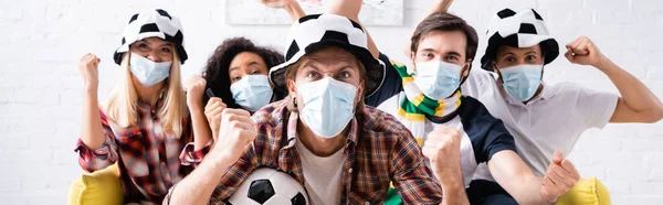 Amigos multiculturais animados em fãs de futebol chapéus e máscaras médicas mostrando gesto de vitória enquanto assiste campeonato, banner — Fotografia de Stock