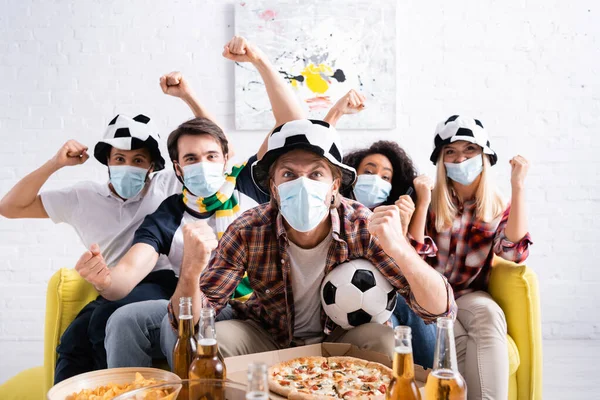 Amigos multiétnicos emocionados en máscaras médicas mostrando gesto ganador mientras ve el campeonato de fútbol en casa - foto de stock