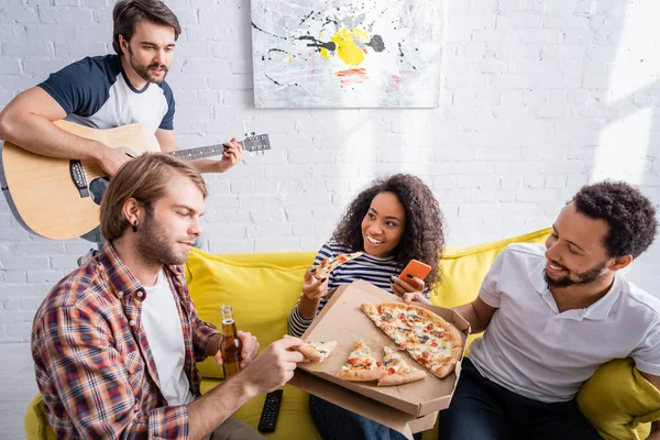 Multikulti-Freunde essen Pizza auf Sofa, während junger Mann Akustikgitarre spielt — Stockfoto