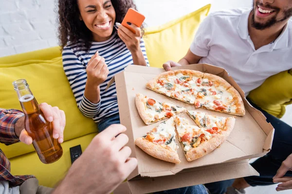 Amigos multiculturales sosteniendo pizza cerca de mujer afroamericana riendo sobre fondo borroso - foto de stock