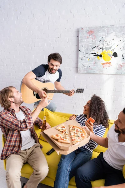 Збуджений чоловік грає на гітарі з веселими багатоетнічними друзями, сидячи на дивані під час вечірки — стокове фото